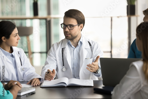 Serious male chief doctor in glasses and white coat speaking at meeting with diverse staff, talking to multiethnic professional colleagues, discussing healthcare job on medical seminar