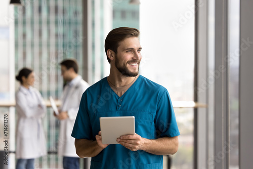 Happy handsome doctor man in blue uniform holding digital tablet, looking away, thinking, smiling, laughing, standing in clinic hall space, using work application, electronic online communication