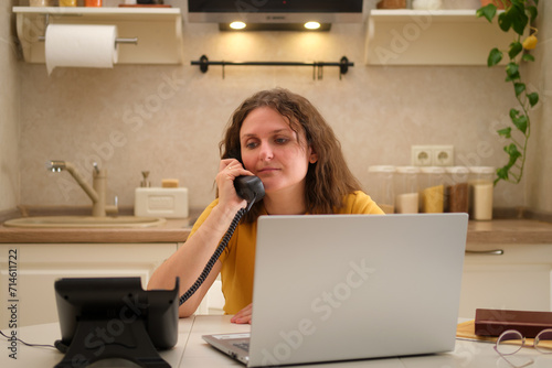 A woman with a laptop is talking on a landline phone at a table in a home kitchen. An adult female businesswoman works from home, a remote office photo