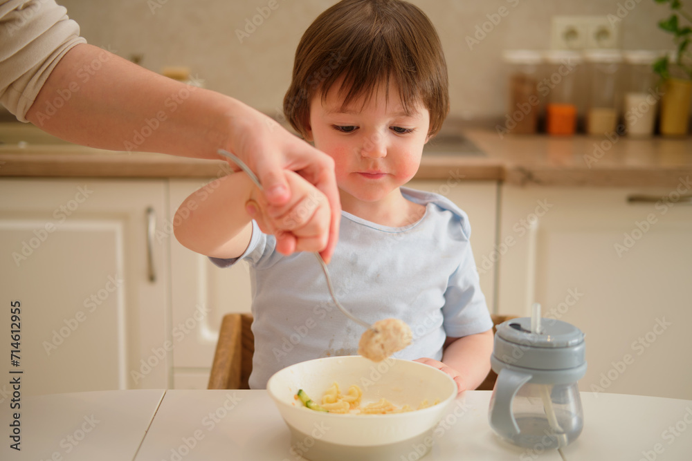 Mother woman teaches toddler baby to eat with a fork in home kitchen. Mom and son are cooking together in the kitchen. Toddler baby two years old at dinner Mother woman teaches toddler baby to eat with a fork in home kitchen. Mom and son are cooking together in the kitchen. Toddler baby two years old at dinner