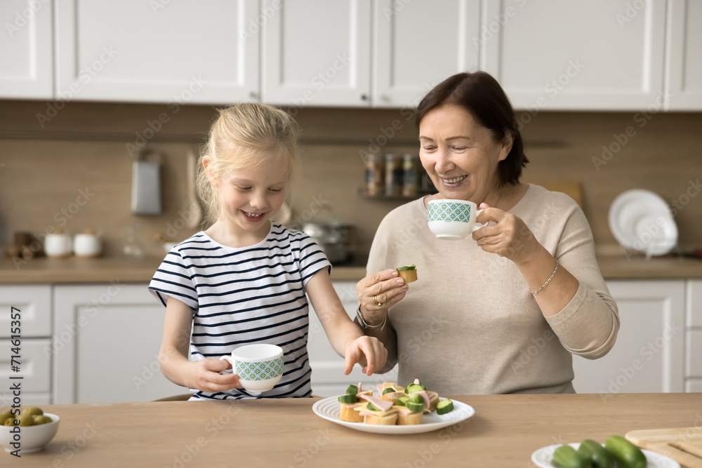 Cheerful granddaughter girl and grandma eating homemade sandwiches in kitchen together, laughing, drinking hot beverage, tea, cocoa, laughing, talking, having breakfast, lunch Cheerful granddaughter girl and grandma eating homemade sandwiches in kitchen together, laughing, drinking hot beverage, tea, cocoa, laughing, talking, having breakfast, lunch