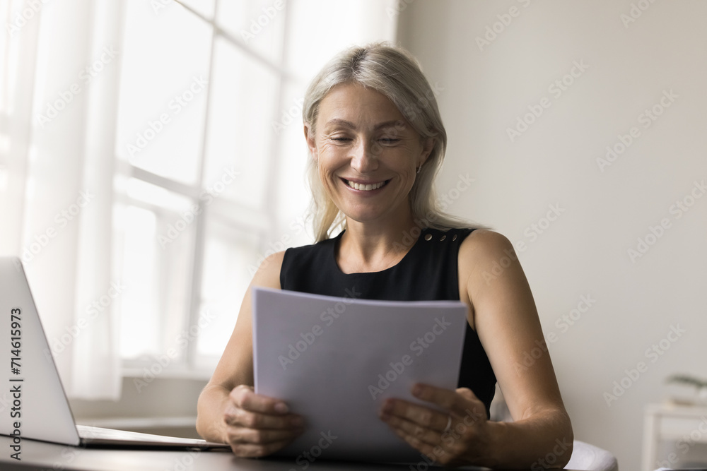 Attractive middle-aged woman sit at desk, hold paper letter, bank statement or notification, do paperwork, satisfied with financial report, get good news, feel happy, loan approval, investment income Attractive middle-aged woman sit at desk, hold paper letter, bank statement or notification, do paperwork, satisfied with financial report, get good news, feel happy, loan approval, investment income