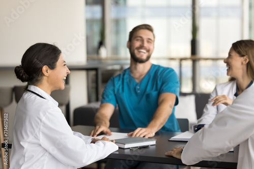 Cheerful young Indian doctor woman laughing at professional meeting with practitioner colleagues, sitting at table, having fun, enjoying leisure, work break, discussing medical job success