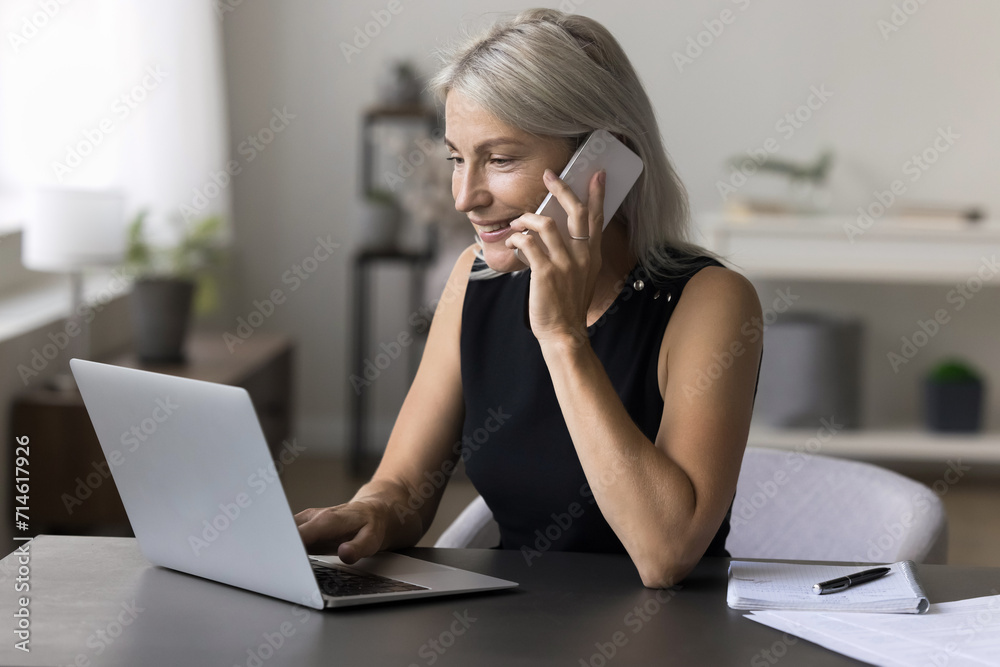 Attractive smiling business woman sit at desk in modern office, hold smartphone, make business call, enjoy pleasant conversation, consulting client by phone, discuss project with partners use laptop