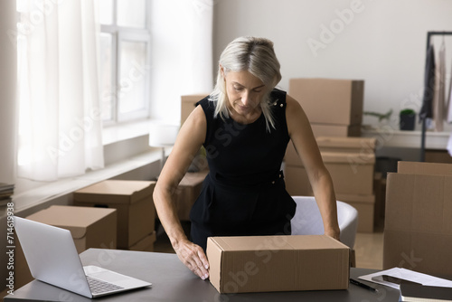 Mature 50s businesswoman packing cardboard box, prepare parcel for dispatch to client, sell goods to customer, ships the items use reliable transport company services. Shipment, distribution, business
