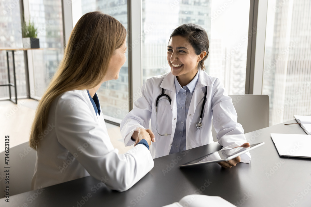 Cheerful diverse clinic colleagues women shaking hands at workplace table, smiling, laughing. Young practitioner giving handshake to happy medical specialist with gratitude, thanking for help