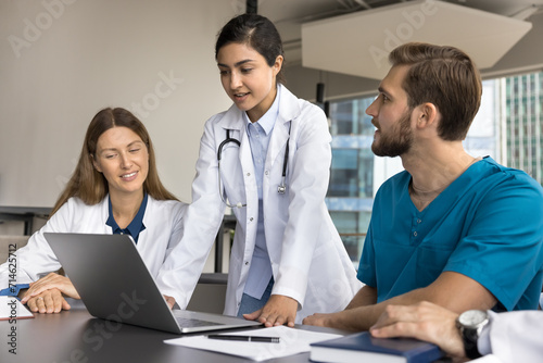 Young Indian medical specialist explaining examination electronic results to colleagues at clinic office table, speaking at laptop. Diverse team of doctors discussing case at computer