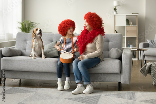 Cheerful little kid girl and grandma in red wigs sitting on couch at trick dog, playing toy drum, singing to music, smiling, laughing, having fun with pet at home masquerade party