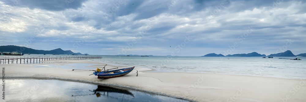 Tranquil beach scene with a solitary wooden boat on white sand, a long pier, and distant mountains under a cloudy sky