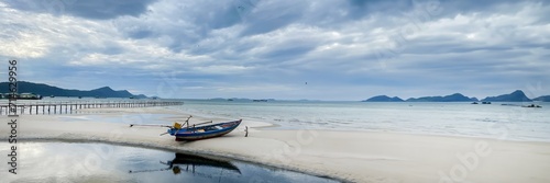 Tranquil beach scene with a solitary wooden boat on white sand, a long pier, and distant mountains under a cloudy sky