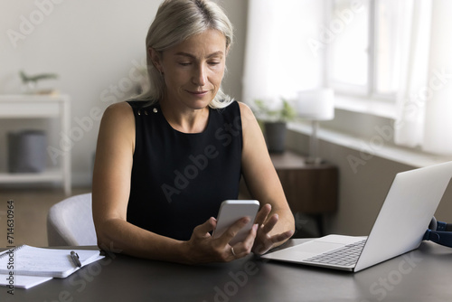 Attractive mature woman sitting at desk hold smartphone, enjoy chat online with colleagues, share messages, send sms use modern technology, browse important information, check schedule, solve business