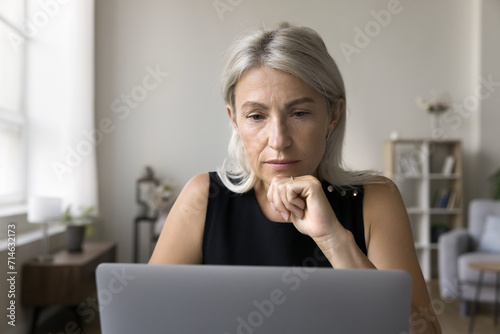 Close up of mature attractive thoughtful businesswoman learn new software, professional program, read received e-mail from client working online using laptop at home office room. Modern tech, business