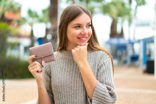 Young pretty blonde woman holding a wallet at outdoors thinking an idea and looking side