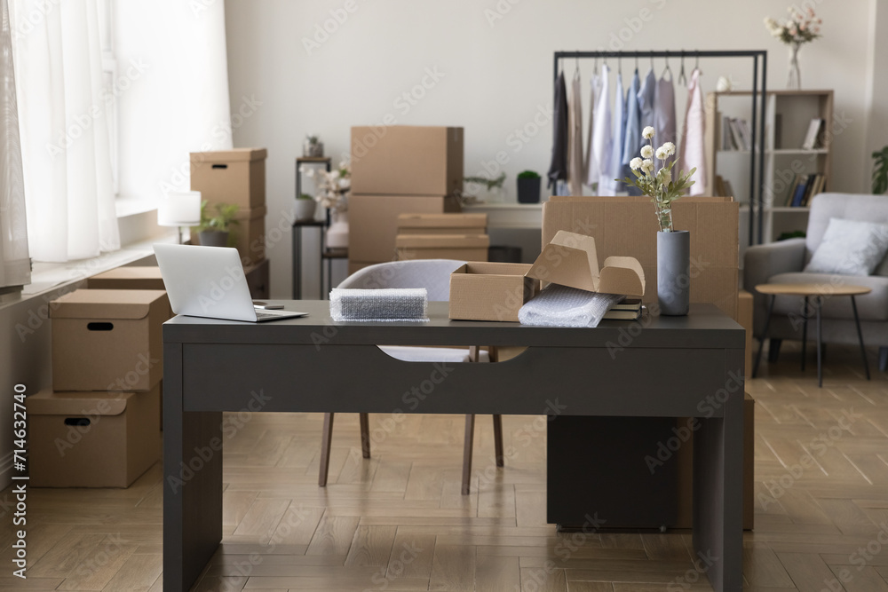 Cozy workplace with desk and stacked heap of cardboard boxes, hanger with clothes, no people. Moving day to new office, workspace of designer, selling fashion attire through internet, small business Cozy workplace with desk and stacked heap of cardboard boxes, hanger with clothes, no people. Moving day to new office, workspace of designer, selling fashion attire through internet, small business
