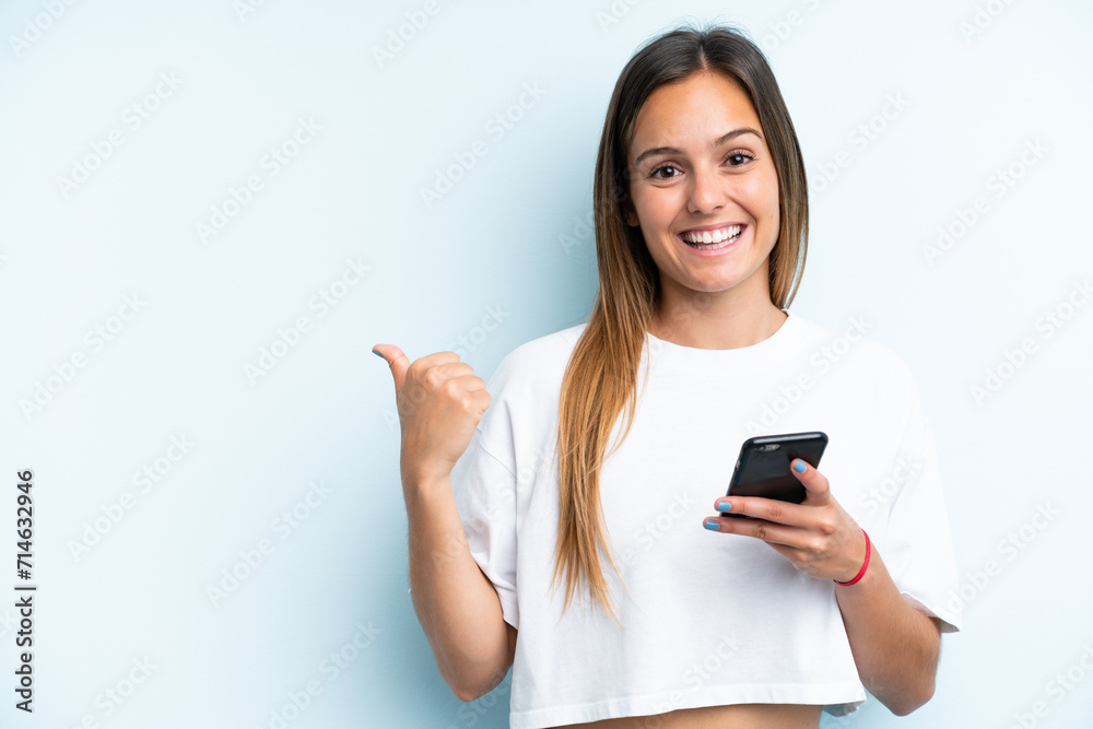 Young caucasian woman isolated on blue background using mobile phone and pointing to the lateral Young caucasian woman isolated on blue background using mobile phone and pointing to the lateral