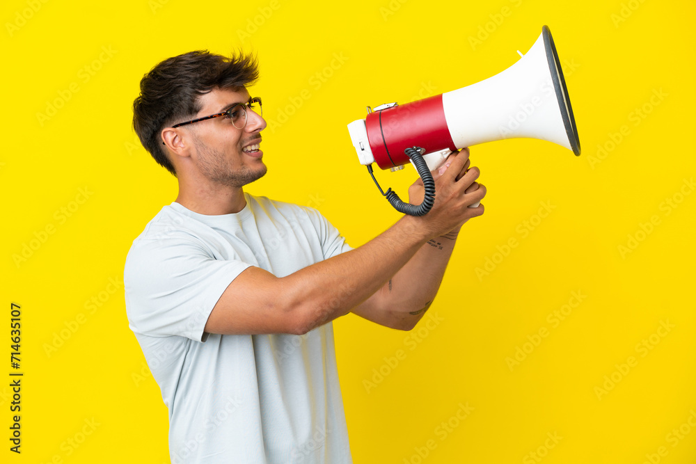 Young caucasian handsome man isolated on yellow background shouting through a megaphone to announce something - obrazy, fototapety, plakaty 