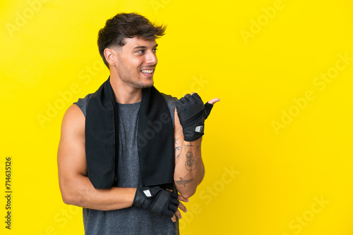 Young sport caucasian man with towel isolated on yellow background pointing to the side to present a product