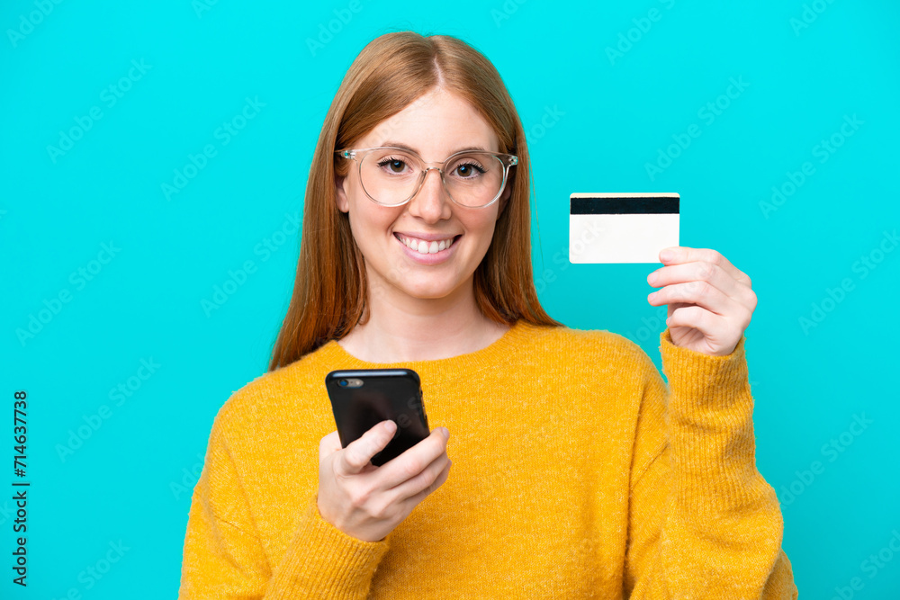 Young redhead woman isolated on blue background buying with the mobile with a credit card Young redhead woman isolated on blue background buying with the mobile with a credit card