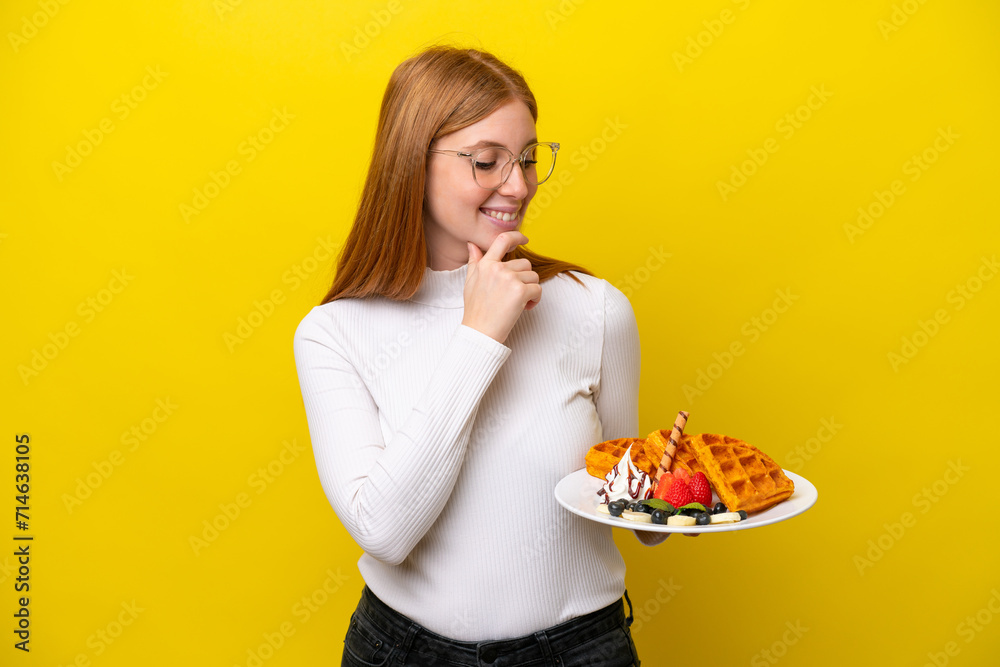 Young redhead woman holding waffles isolated on yellow background looking to the side and smiling