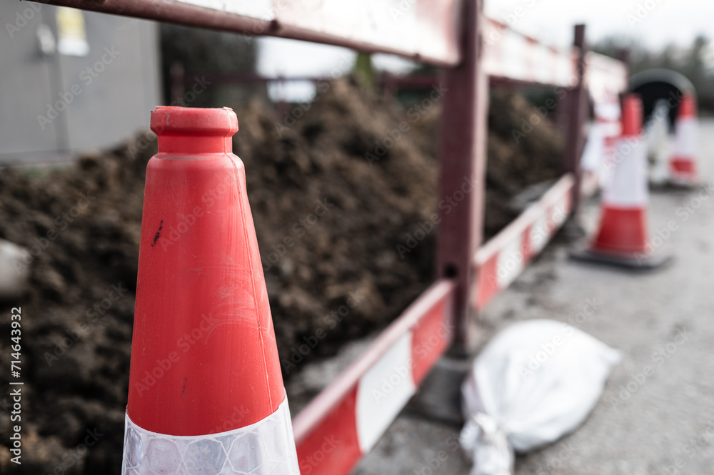 Shallow focus of a standard British traffic cone seen at the edge of a roadworks barrier. The soil heap can be seen, as new gas pipes are being laid adjacent to the road.
