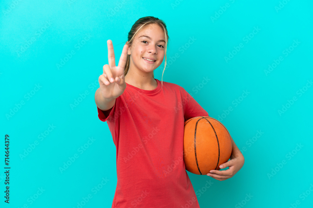 Little caucasian girl playing basketball isolated on blue background smiling and showing victory sign