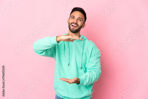 Young caucasian man isolated on pink background holding copyspace imaginary on the palm to insert an ad