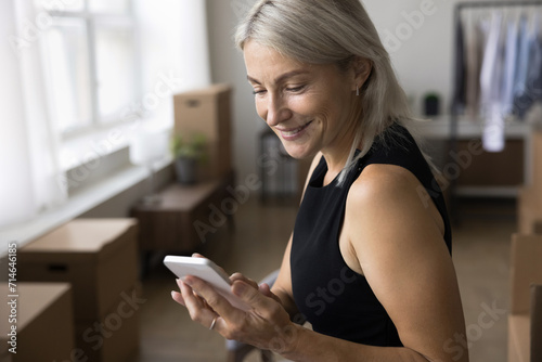 Close up shot of smiling mature 45s woman using smartphone working in warehouse, make call to delivery services, reliable transporting company on relocation day, stacked cardboard boxes on background photo