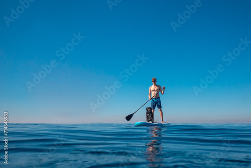 A man paddles on a Stand Up Paddle, SUP board with a black Brittany spaniel dog on the sea. Active lifestyle summer vacation concept