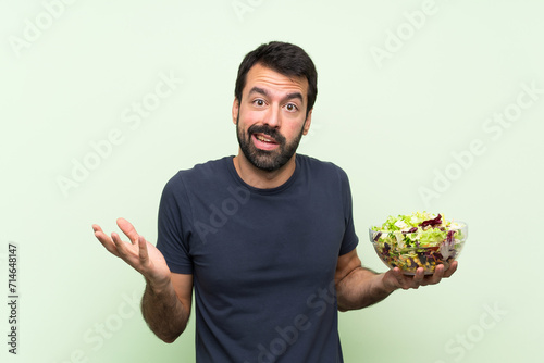 Young handsome man with salad over isolated green wall making doubts gesture