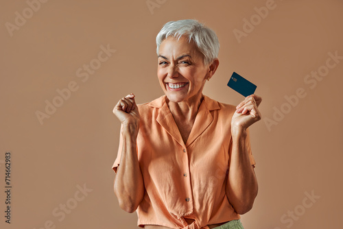Smiling happy sincere emotional crafty satisfied mature beautiful gray haired woman in pastel orange shirt holding credit card and sincerely happy standing on beige background.