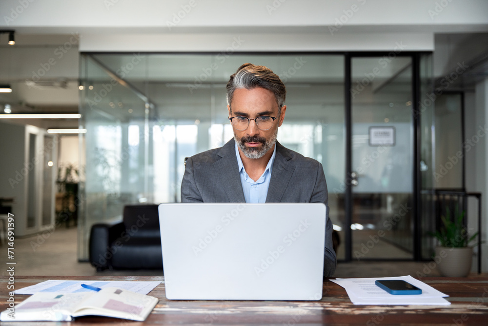 Mature Indian or Latin business man ceo trader using computer, typing, working in modern office, doing online data market analysis, thinking planning tech strategy looking at laptop with copy space. Mature Indian or Latin business man ceo trader using computer, typing, working in modern office, doing online data market analysis, thinking planning tech strategy looking at laptop with copy space.