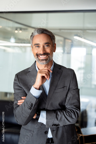 Vertical closeup portrait of handsome hispanic senior business man smiling at camera. European latin confident mature good looking middle age leader male businessman standing on blur office background