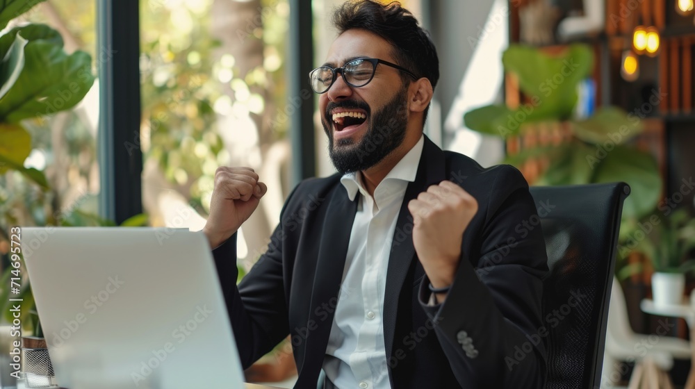 Excited indian or arabian businessman in suit, sits in a modern office, looking at laptop, happy by good news, rejoice by win, raising hand in yes gesture, celebrating project success, big profit Excited indian or arabian businessman in suit, sits in a modern office, looking at laptop, happy by good news, rejoice by win, raising hand in yes gesture, celebrating project success, big profit