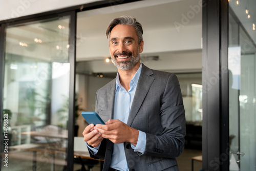Portrait of happy smiling mature European latin businessman holding smartphone in office, copy space. Middle aged man using cell phone mobile app and looking at camera. Applications for business.