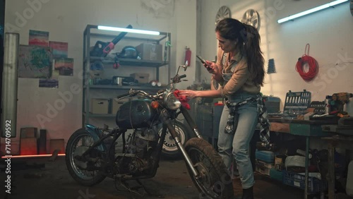 Young woman wipes vintage retro motorcycle and taking photo on smartphone. Young repair woman showing condition of repaired motorbike to customer through video call. Maintenance and repairing concept. photo