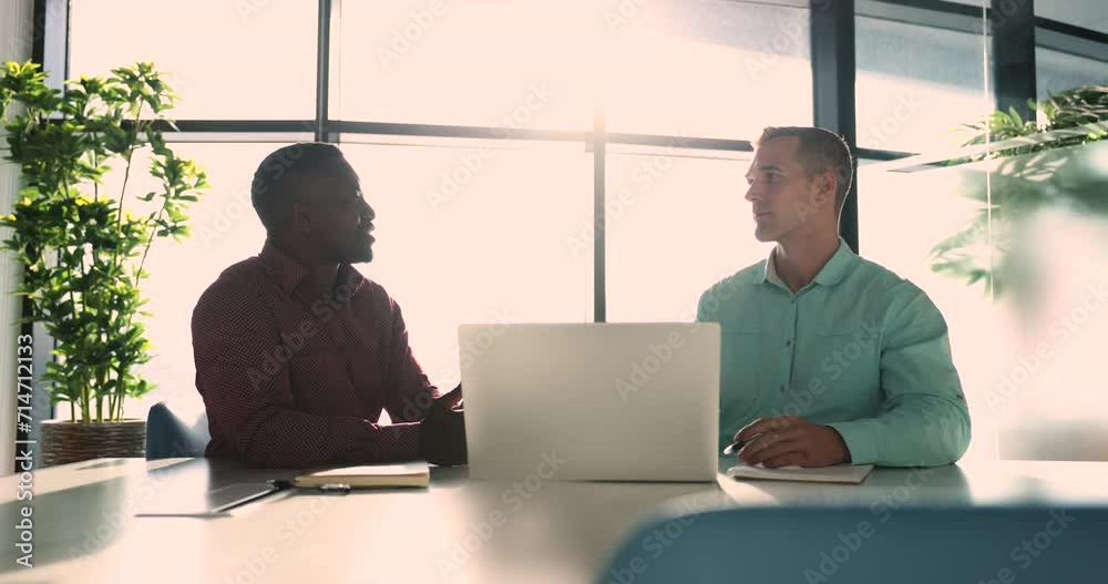 Two multi-ethnic men, teammates consider joint task, working sit at desk with modern laptop, hold discussion, analyzing creative ideas, planning start-up strategy, search solution engaged in teamwork Two multi-ethnic men, teammates consider joint task, working sit at desk with modern laptop, hold discussion, analyzing creative ideas, planning start-up strategy, search solution engaged in teamwork