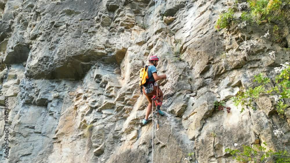 Male climber carving a new climbing route with using rock drills and rotary hammers on the climbing rope. Climber wears a red helmet and a yellow backpack. Male climber carving a new climbing route with using rock drills and rotary hammers on the climbing rope. Climber wears a red helmet and a yellow backpack.
