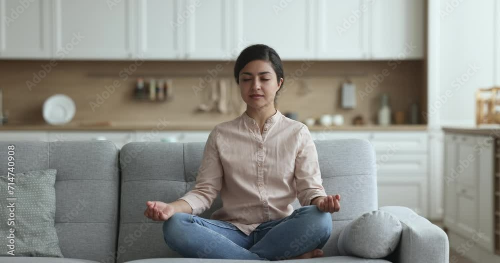 Calm peaceful young Indian yogi girl meditating on home sofa, sitting in yoga pose with closed eyes, keeping zen mudra, calm hand on knees gesture, taking deep breath, smiling Calm peaceful young Indian yogi girl meditating on home sofa, sitting in yoga pose with closed eyes, keeping zen mudra, calm hand on knees gesture, taking deep breath, smiling