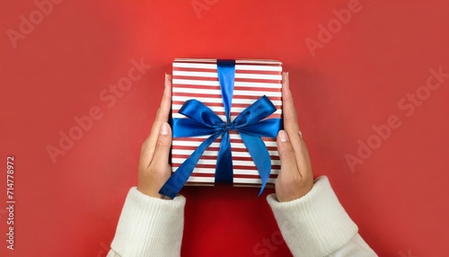 female s hands holding striped gift box with blue ribbon on red background christmas new year valentine s day and birthday concept