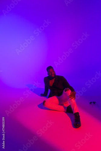 A young man in a jacket and white trousers sits on the floor in neon light.