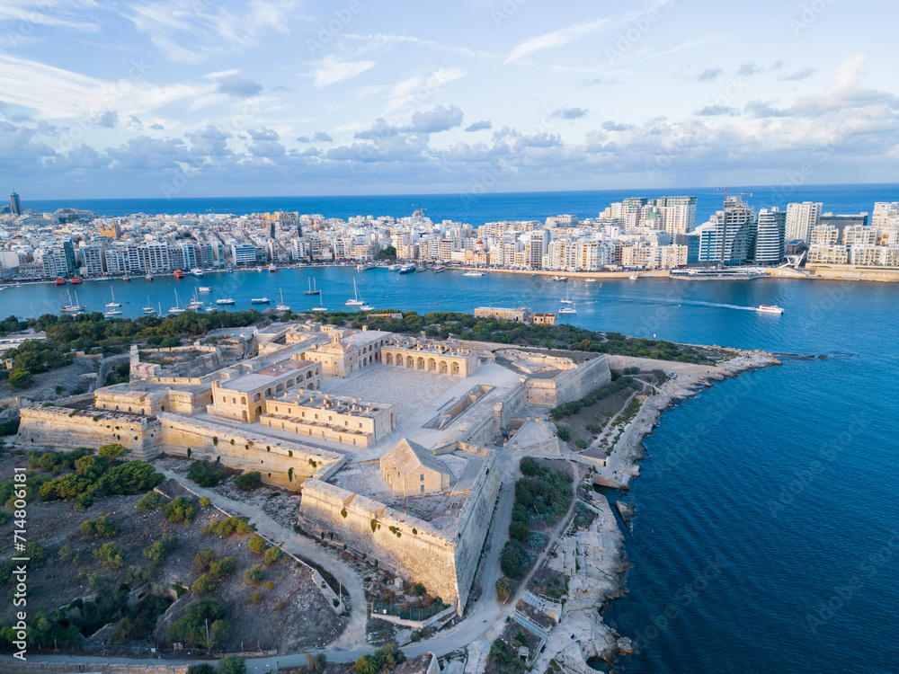 Fort Manoel at sunset with Silema skyline. Higher altitude aerial view on fort and harbor at golden hour. Natural blue and golden colors with clouds in the sky.