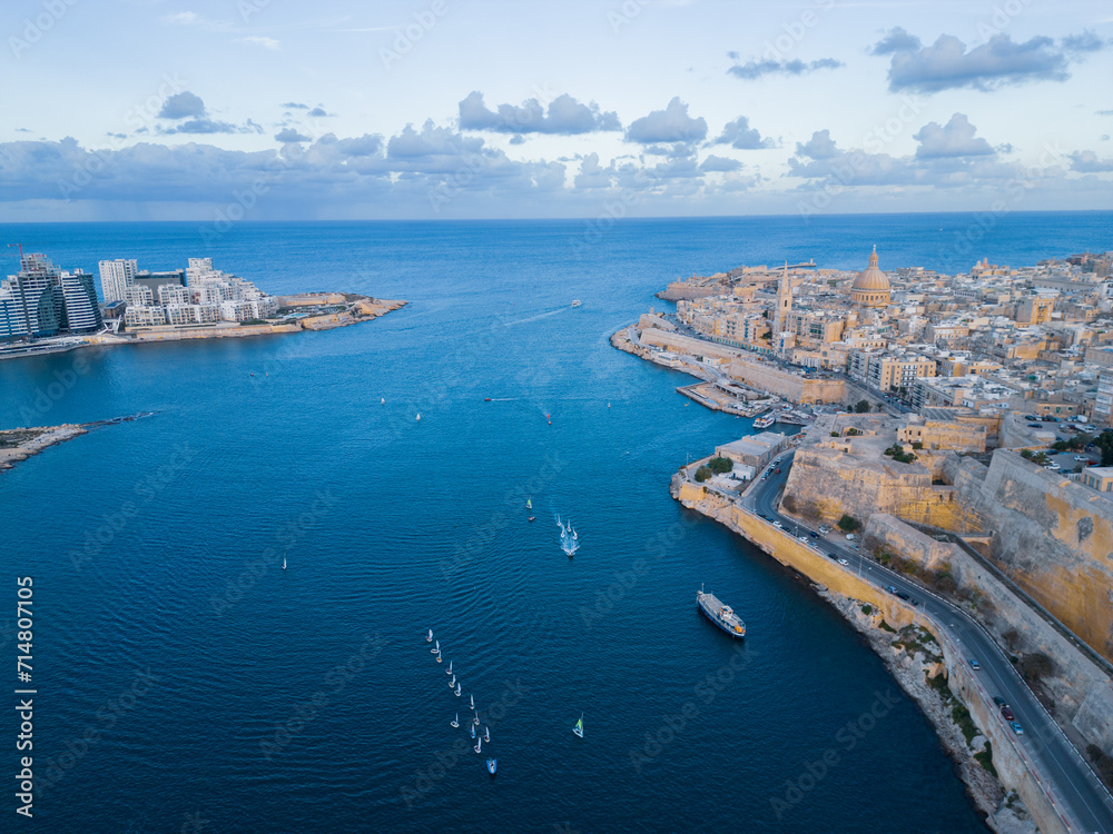 Historical Valletta and modern Sliema natural harbor. High altitude aerial view at sunset before dusk. Sailing boats returning into harbor. Clear blue sky with clouds.