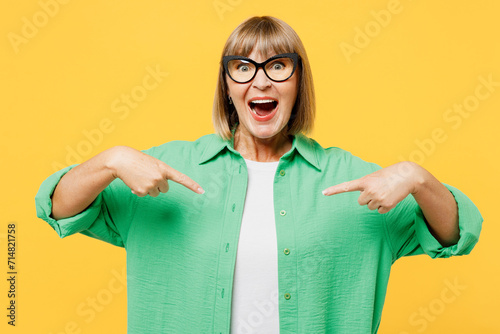Elderly excited happy fun blonde woman 50s years old she wear green shirt glasses casual clothes point index fingers on herself isolated on plain yellow background studio portrait. Lifestyle concept. photo