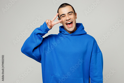 Young cheerful happy smiling satisfied fun middle eastern man he wear blue hoody casual clothes cover eye with victory sign isolated on plain solid white background studio portrait. Lifestyle concept.