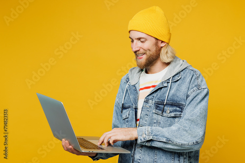 Side view young blond IT man wears denim shirt hoody beanie hat casual clothes hold use work on laptop pc computer typing message isolated on plain yellow background studio portrait Lifestyle concept © ViDi Studio