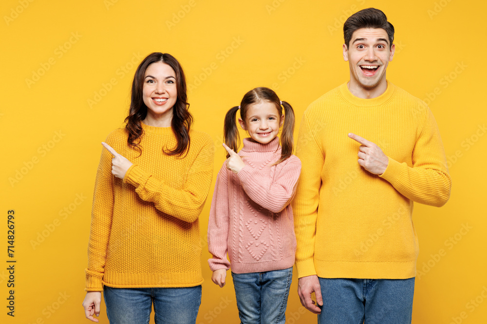 Fototapeta premium Young happy parents mom dad with child kid girl 7-8 years old wear pink knitted sweater casual clothes point index finger aside on empty area isolated on plain yellow background. Family day concept.