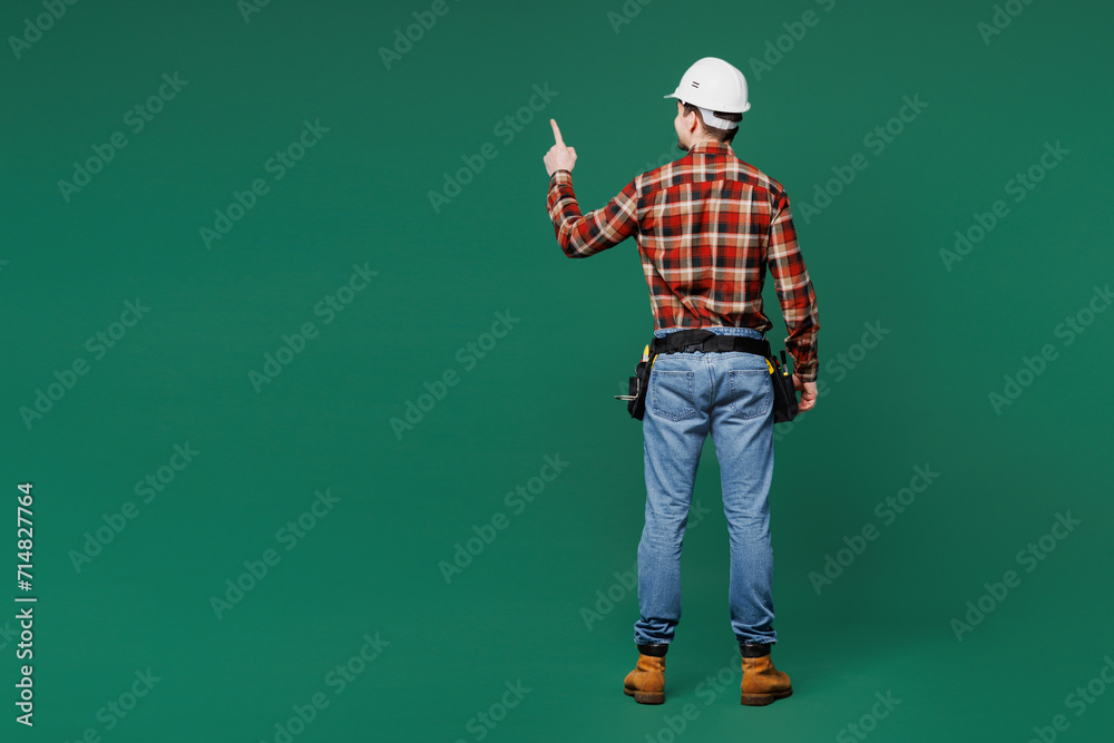 Full body back view young laborer handyman man wear red shirt hardhat point index finger aside on area isolated on plain green background Instruments for renovation apartment room Repair home concept Full body back view young laborer handyman man wear red shirt hardhat point index finger aside on area isolated on plain green background Instruments for renovation apartment room Repair home concept