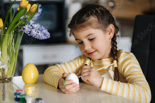 Adorable little girl painting eggs on kitchen at home. Cute girl in apron. Background of easter decoration