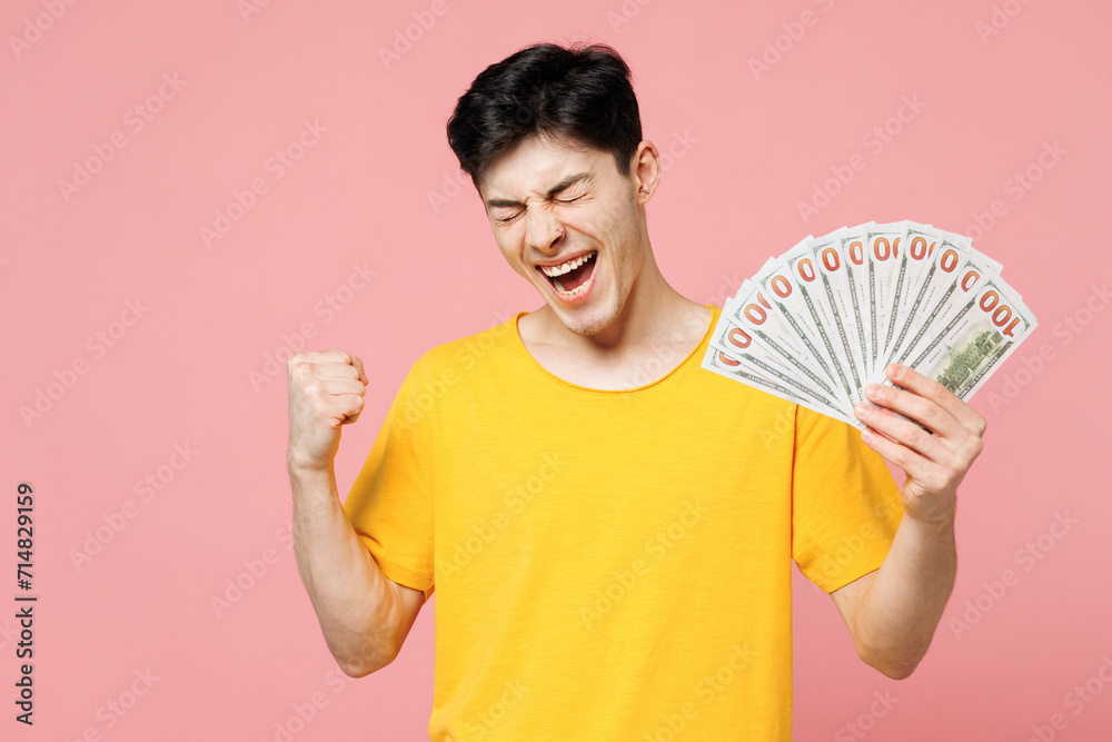 Young smiling Caucasian man wears yellow t-shirt casual clothes hold in hand fan of cash money in dollar banknotes do winner gesture isolated on plain pastel light pink background. Lifestyle concept. Young smiling Caucasian man wears yellow t-shirt casual clothes hold in hand fan of cash money in dollar banknotes do winner gesture isolated on plain pastel light pink background. Lifestyle concept.