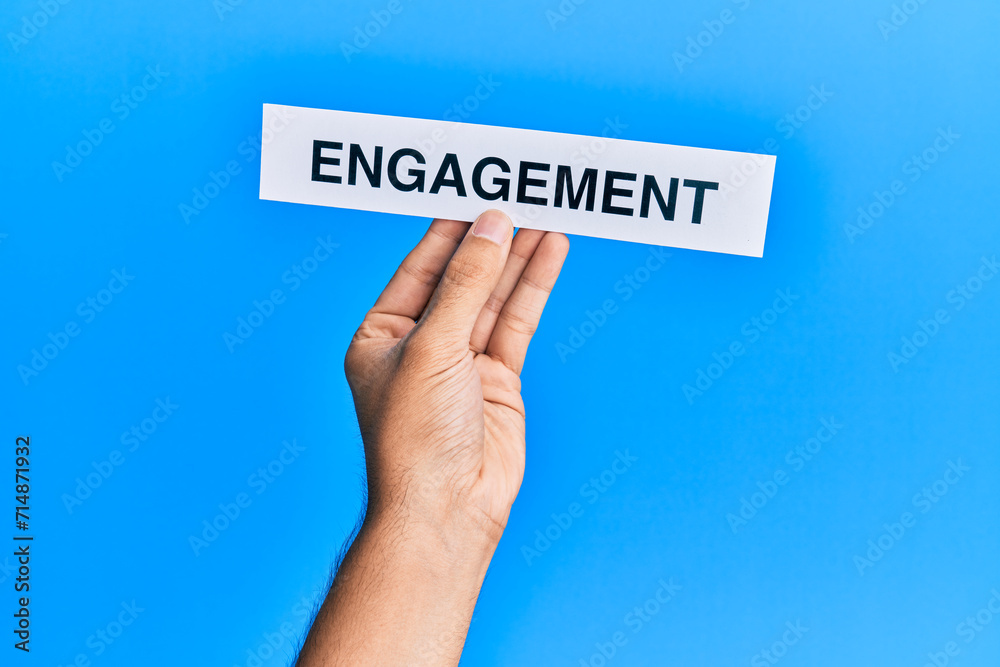 Hand of caucasian man holding paper with engagement word over isolated blue background Hand of caucasian man holding paper with engagement word over isolated blue background
